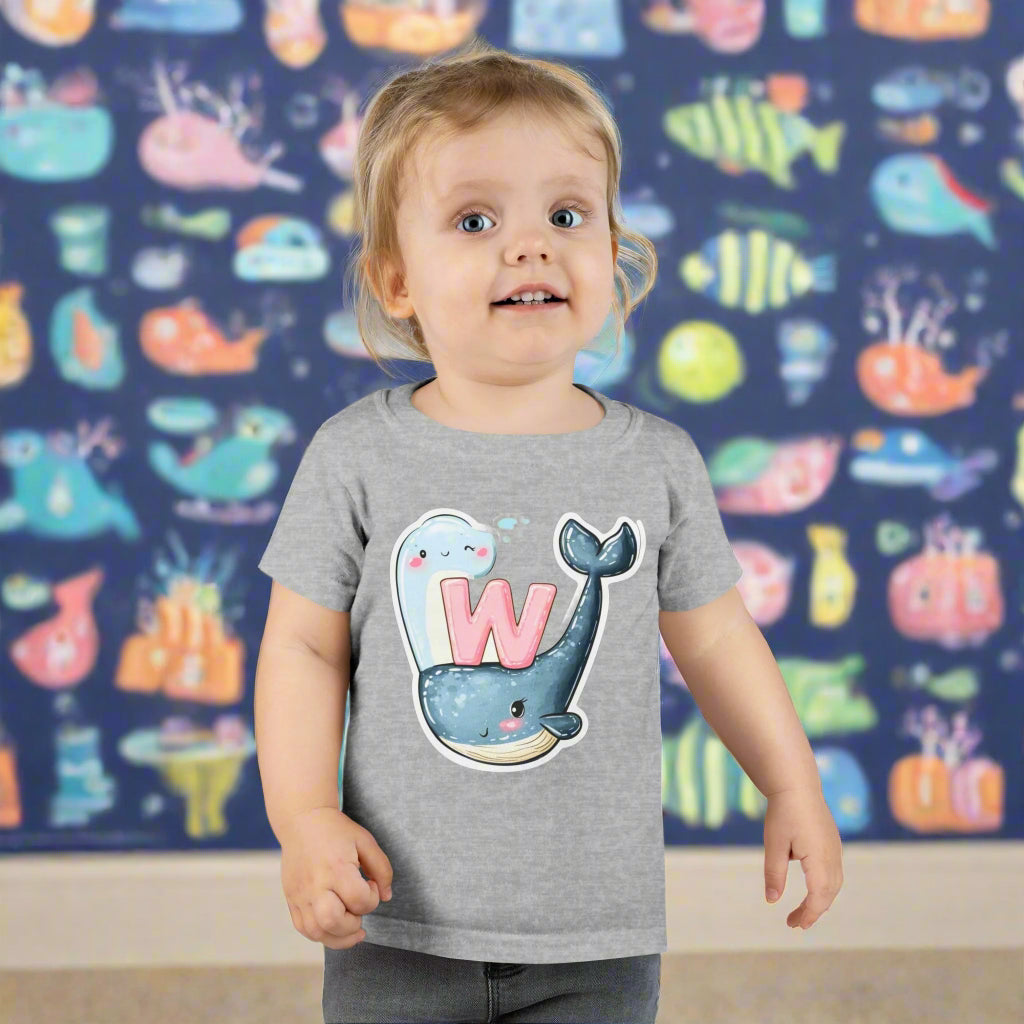 A toddler wearing a gray T-shirt with a whimsical whale design and the letter 'W'.
