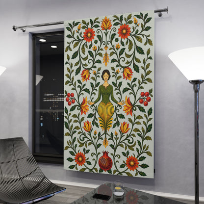 A large fabric wall hanging or tapestry displayed in a modern room. The tapestry features a folk-art style design with a central female figure surrounded by symmetrical floral and vine motifs in green, red, orange, and yellow on a light background.